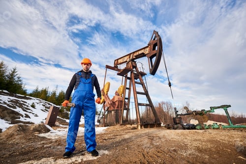 Preview: Oil worker in uniform and helmet working in an oilfield next to a pump jack on a sunny day