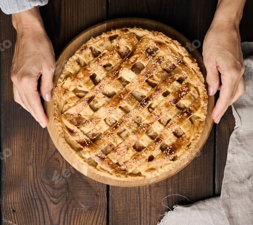 Preview: Two female hands hold a round baked pie with apple filling on a wooden board, brown table.