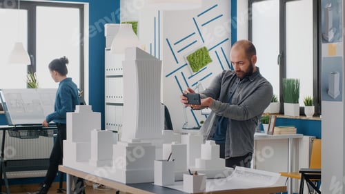 Preview: Man engineer using smartphone to work on building model and layout in architectural office