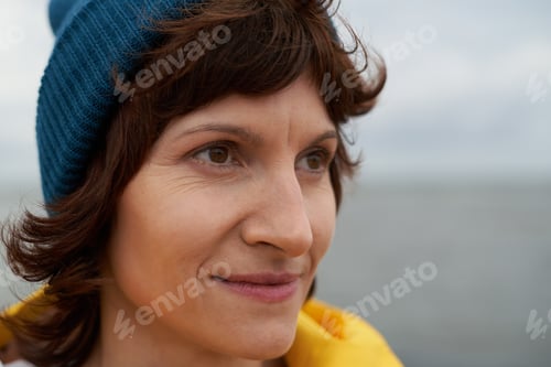 Preview: Crop closeup portrait of mature beautiful woman looking away in bright yellow raincoat and blue hat
