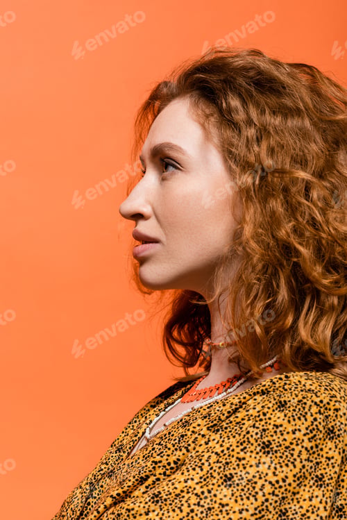 Preview: Side view of stylish redhead woman in necklaces and yellow blouse with abstract pattern looking away