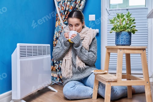 Preview: Teen female in sweater scarf warming near electric heating radiator, drinking hot drink