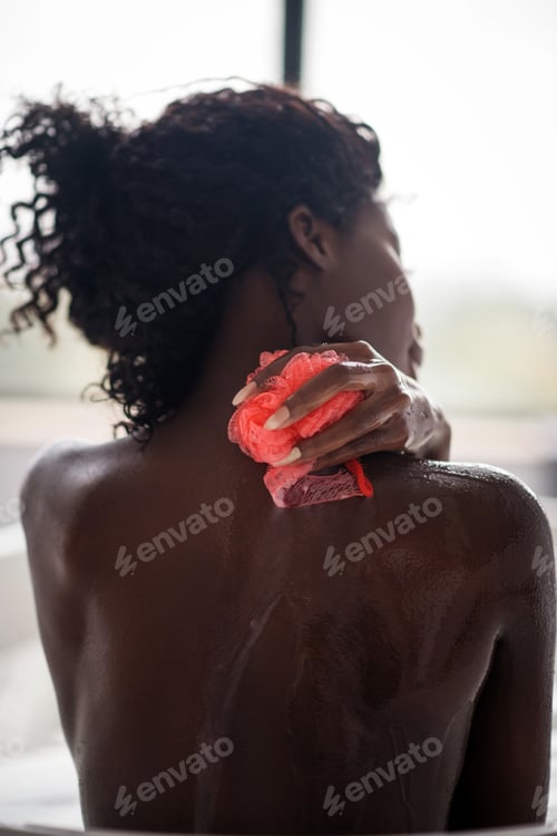 Preview: Curly dark-haired woman using loofa while cleaning her back
