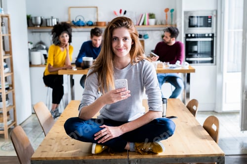 Preview: Woman with cell phone sitting on dining table at home with friends in background