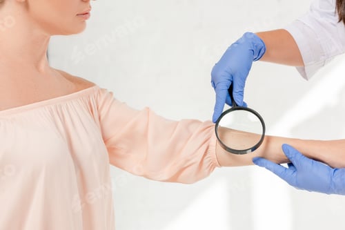 Preview: cropped view of dermatologist examining skin of patient with magnifying glass in clinic