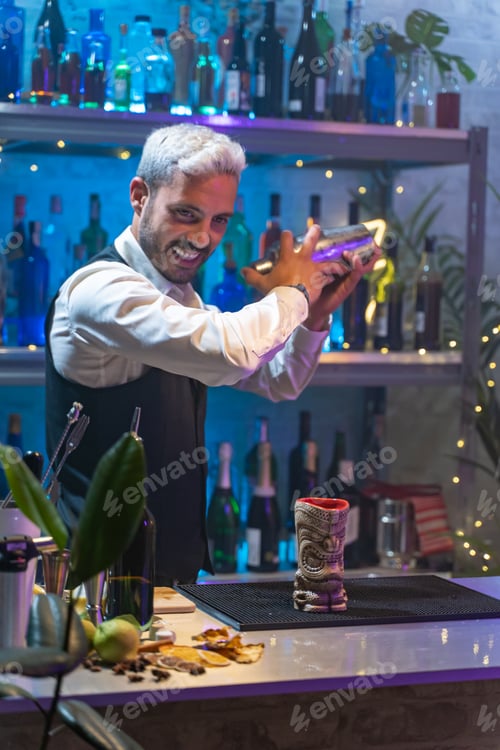 Vista previa: El camarero profesional con camisa blanca y delantal negro sostiene una coctelera de acero en sus manos.