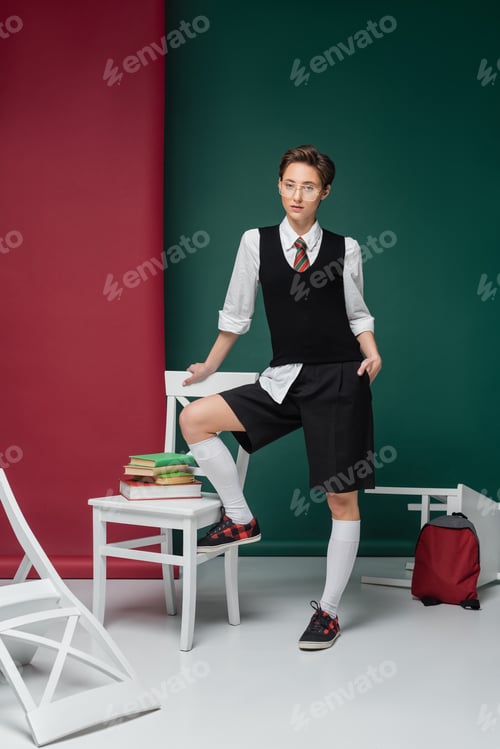 Preview: full length of stylish young student in eyeglasses standing with hand in pocket around chairs with
