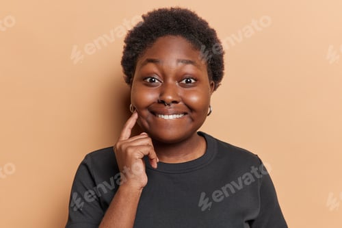 Preview: Portrait of African chubby woman bites lips keeps finger on cheek looks gladfully at camera dressed