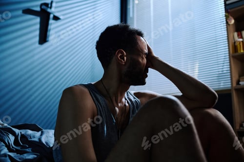 Preview: Young stressed man touching head while sitting by bed after sleepless night