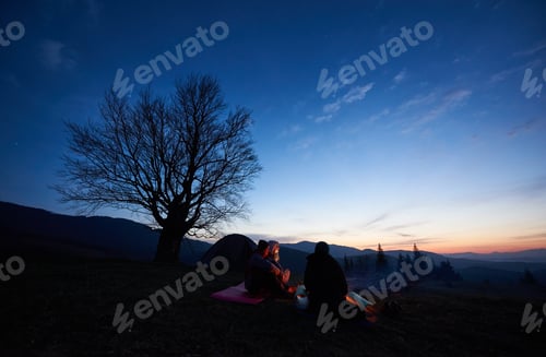 Preview: Camping site in mountains at dawn. Group of three tourists sitting in front of tent