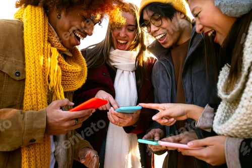 Preview: Multiracial friends looking at phone together on winter day. Young woman pointing at device screen.