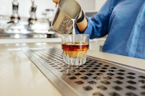 Preview: Unrecognizable barista pouring milk from a stainless steel milk jug into a glass for flat white