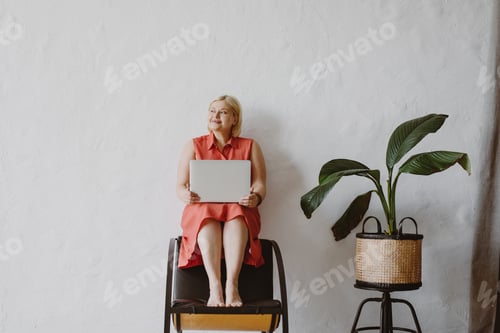 Preview: Young woman sitting on chair using laptop in front of white wall.
