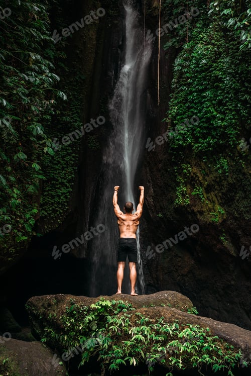 Preview: Back view of a man standing at a waterfall with his hands raised