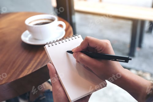 Preview: Closeup image of a woman's hands writing down on a white blank notebook with coffee cup on table