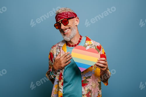 Preview: Joyful hipster senior man in funky shirt holding heart shaped rainbow flag against blue background