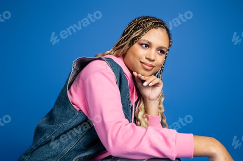 Preview: charming and dreamy dark skinned woman in denim vest and hoodie looking away on blue background