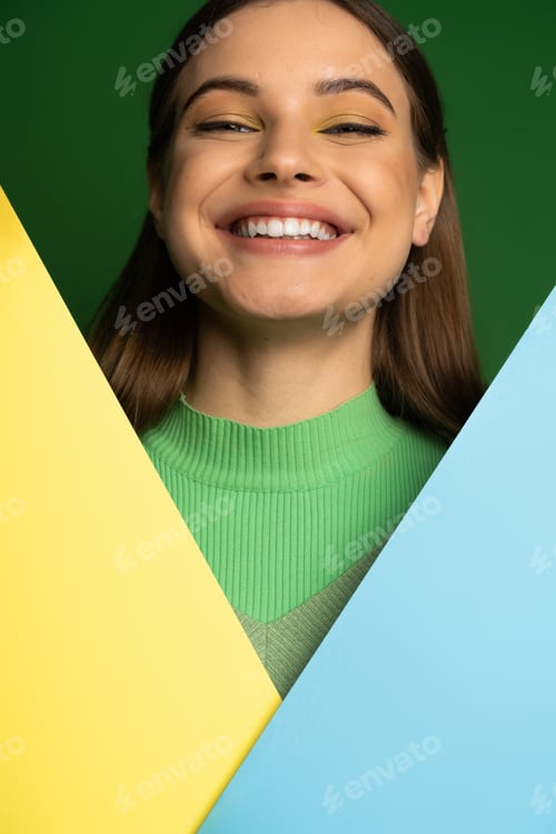 Preview: Positive teen girl with visage looking at camera near colorful paper isolated on green