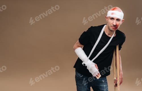 Preview: A battered man with a bandaged head and a cast on his arm stands on crutches on a gray background