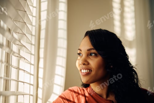 Preview: Happy black woman looking through the window at home.