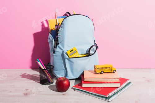 Preview: full backpack with school stationery near toy school bus on books, ripe apple and pen holder on pink
