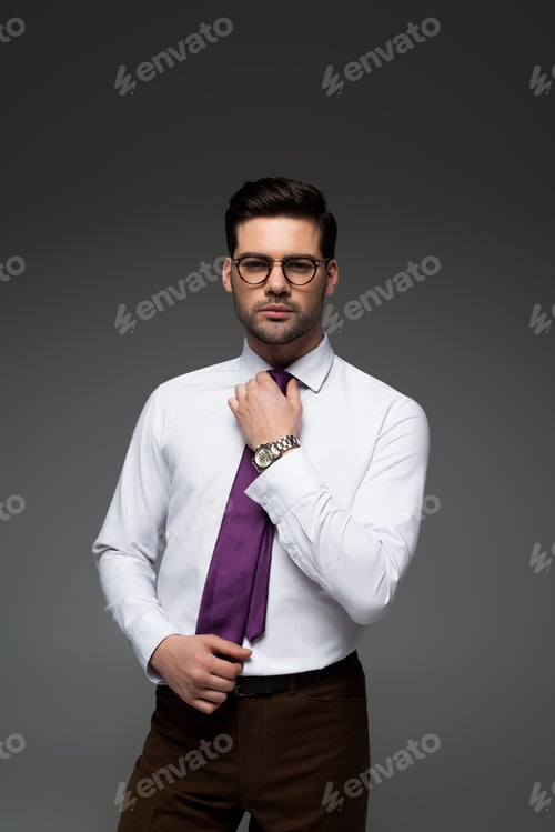 Preview: Young businessman in glasses correcting tie isolated on grey