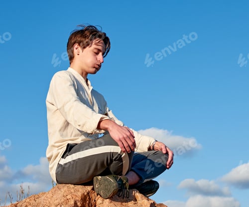 Preview: Shallow focus of a Spanish young male sitting on a rock and meditating against the cloudy blue sky