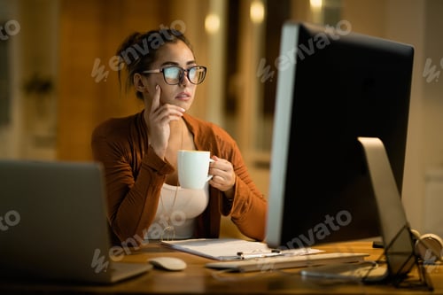 Preview: Young pensive woman drinking tea while reading e-mail on computer at home.