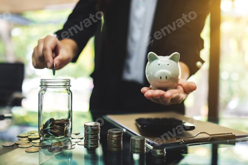 Preview: Businesswoman holding a piggy bank while putting coin into a glass jar