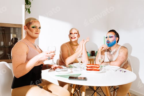 Preview: Group of adult men drag queens in wigs sitting at table having fun and eating cake in daylight