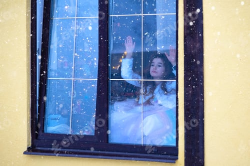 Preview: Girl looks out window at snowfall from Cozy festive home outside with the warm light of fairy lights