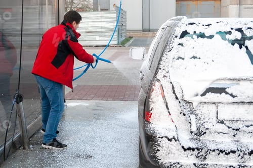 Preview: A man in a red jacket covers a black car with foam at a self-service car wash. Back view