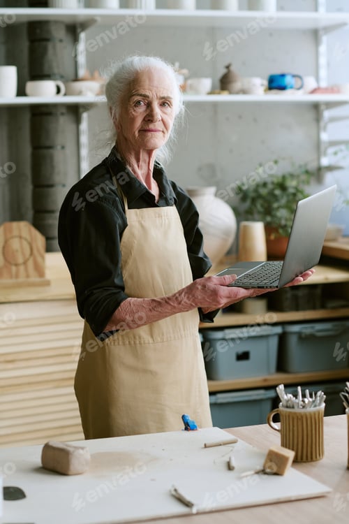 Preview: Senior Woman Holding Laptop While Standing in Pottery Workshop
