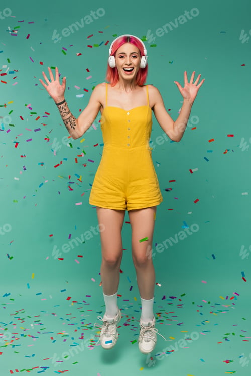 Preview: full length of positive young woman with pink dyed hair in wireless headphones jumping near colorful