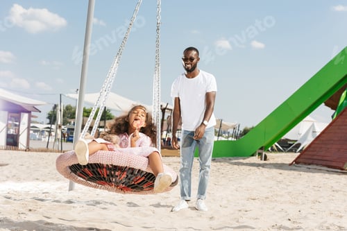 Preview: african american daughter eating lollipop on spider web nest swing at amusement park