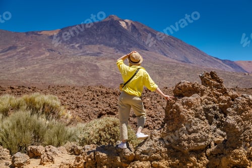 Preview: Back view of a tourist woman in a yellow shirt and straw hat, admiring the grandeur of Mount Teide