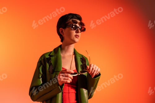 Preview: trendy woman in green leather jacket posing in several sunglasses and looking away on orange
