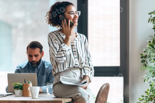 Preview: Smart businesswoman talking with mobile phone while cheeking some documents in modern workspace.