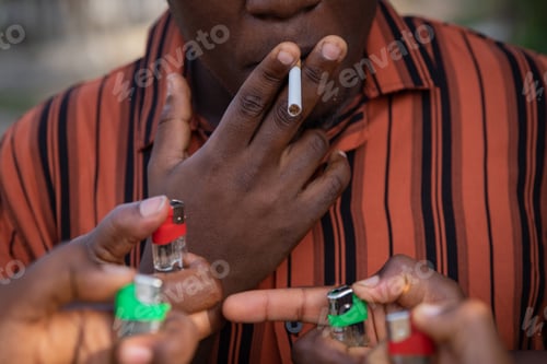 Preview: A boy with a cigarette in his mouth while hands with lighters want to light the cigarette.