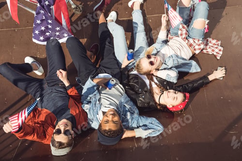 Preview: elevated view of teenagers having fun and lying with american flag in skateboard park, hipster style