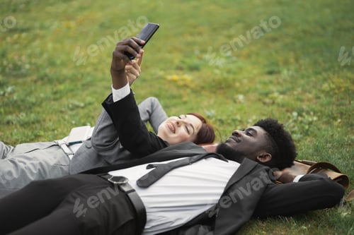 Preview: Multiracial well-dressed young business people resting lying on green grass with smartphone