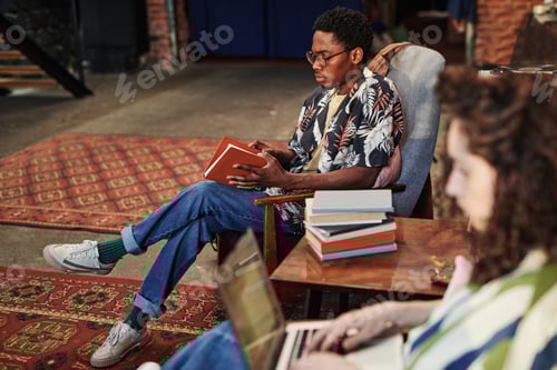 Preview: Serious guy opening book in brown cover while sitting in armchair in living room