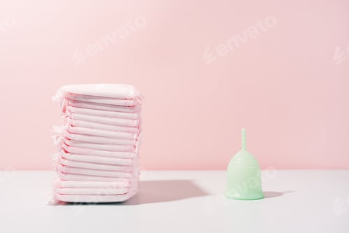 Preview: Menstrual cup next to pack of pads on white table against pink background