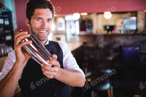 Preview: White mid adult male bartender shaking stainless steel cocktail shaker behind wooden bar counter