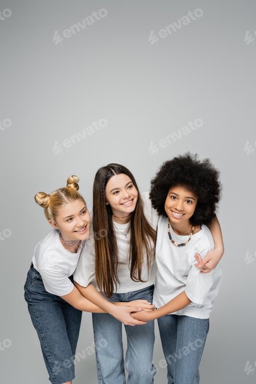 Preview: Cheerful and multiethnic teenage girlfriends in white t-shirts and jeans hugging brunette friend