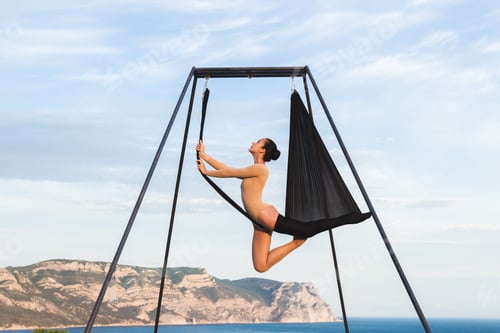 Preview: Woman practicing fly-dance yoga poses in a hammock on the coast with views of the mountains