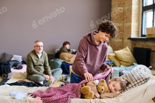 Preview: Young brunette woman covering her sleeping son with teddybear with blanket