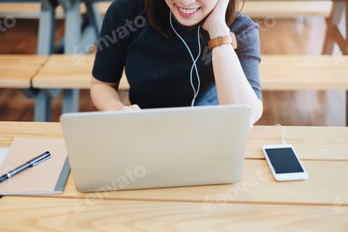 Preview: Young businesswoman playing music with her smartphone and wearing earphones while using laptop in