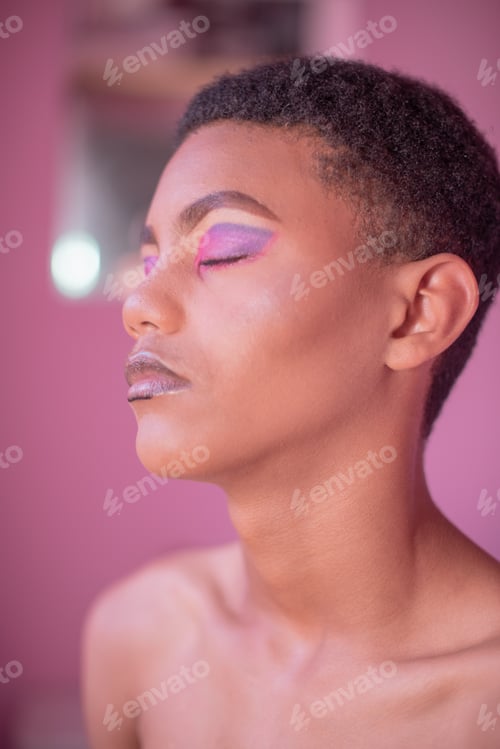 Preview: Portrait of young queer person with make up on against pink background.