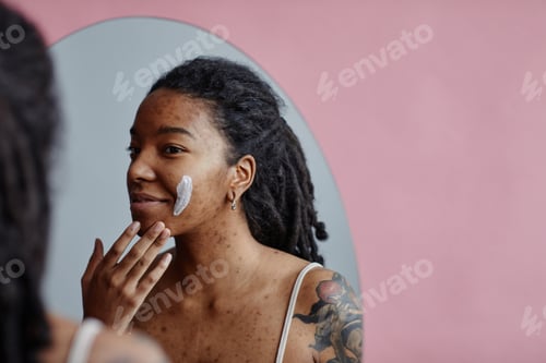 Preview: Young black woman with acne scars using face cream looking in mirror